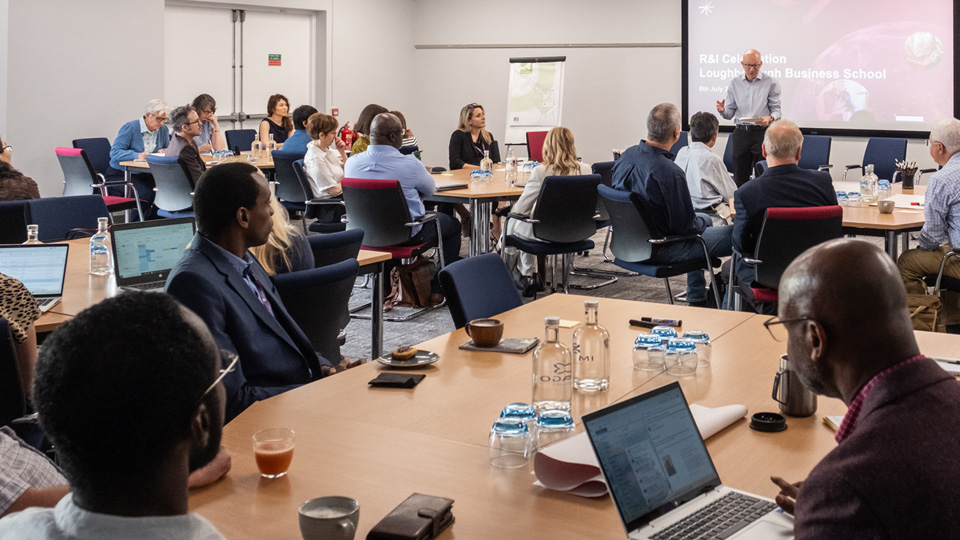 People seated around tables in a conference room during a presentation, with a screen displaying 